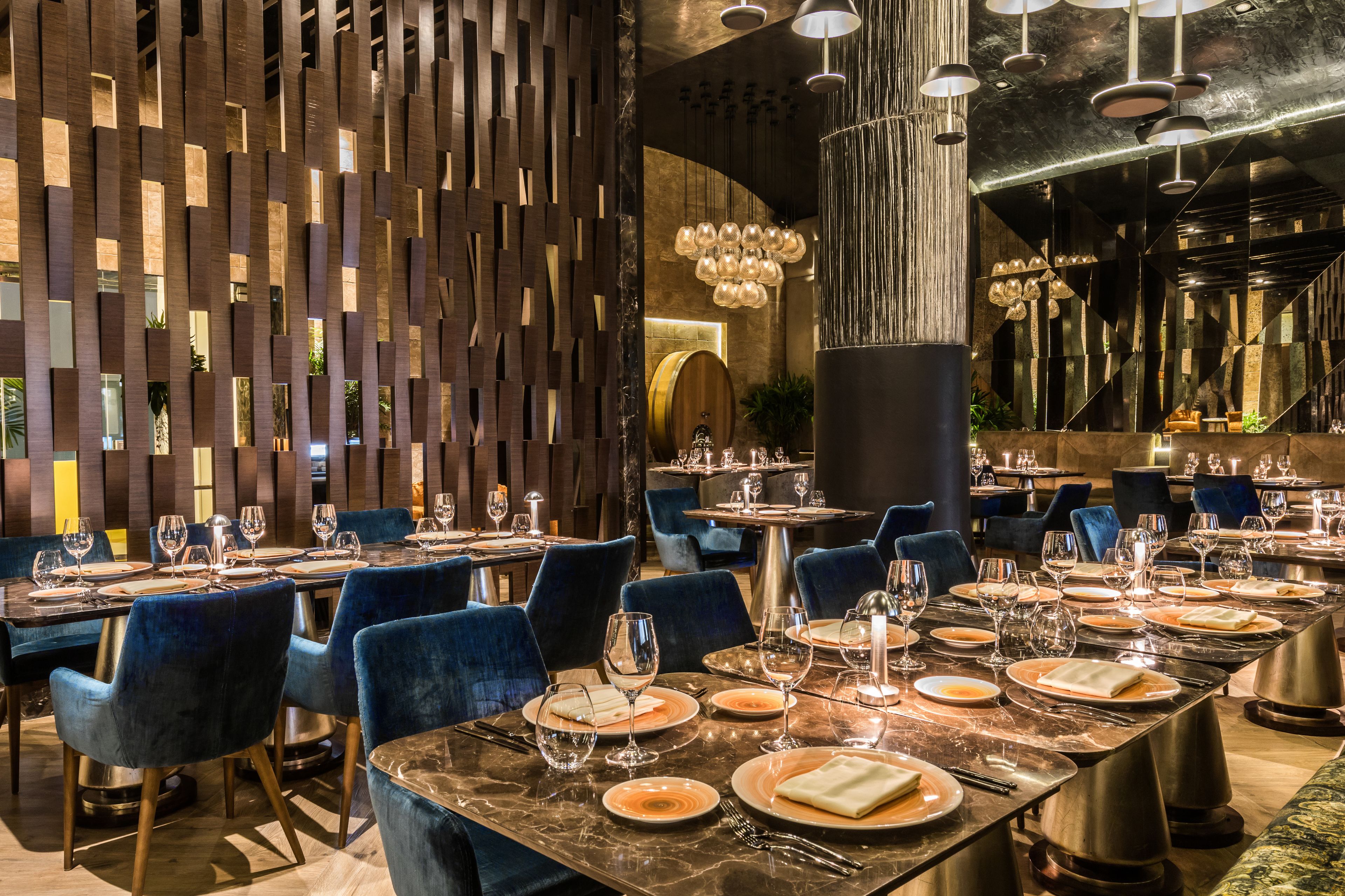 Un restaurant doté de nombreuses tables, de chaises recouvertes d’un tissu bleu et d’un décor intérieur moderne comprenant des panneaux en bois et des luminaires suspendus.