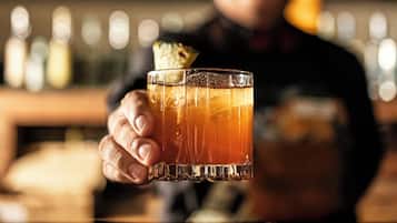 A person holding a glass with a beverage, a bar counter, and a blurred background with lights.