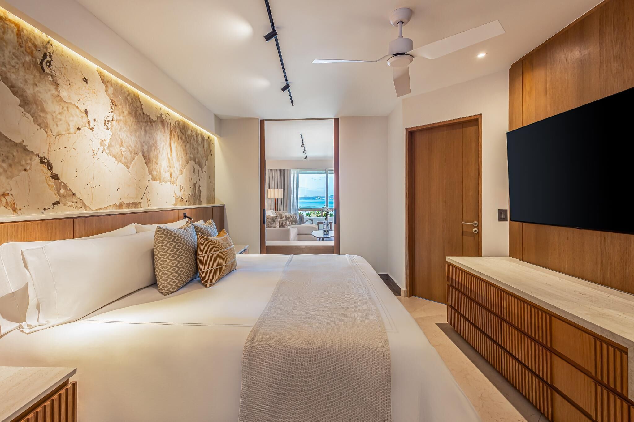 Una habitación de hotel moderna con una cama grande, cabecera de madera, televisor y vista al mar a través de una puerta de cristal.