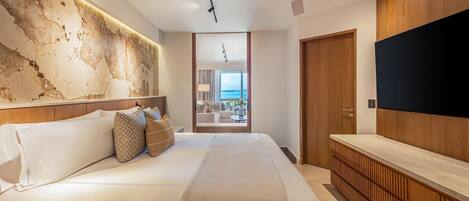 A modern hotel room with a large bed, a wooden headboard, a TV, and a view of the sea through a glass door.