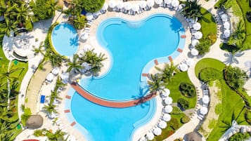 An aerial view of a resort with a large, uniquely shaped swimming pool surrounded by lounge areas with white umbrellas and palm trees.