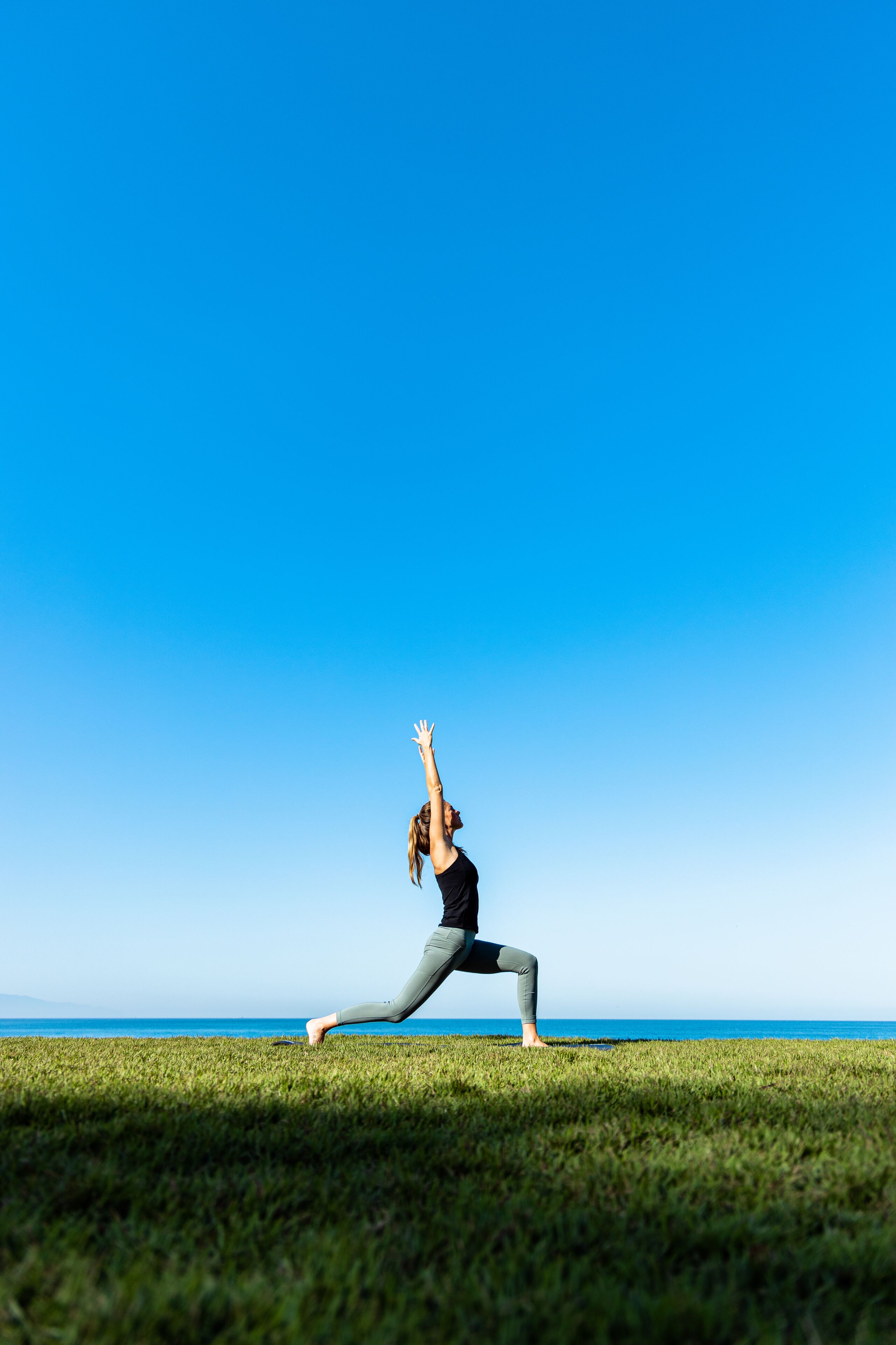 Una persona realizando una postura de yoga en un campo cubierto de césped, con un cielo azul despejado y el océano al fondo.
