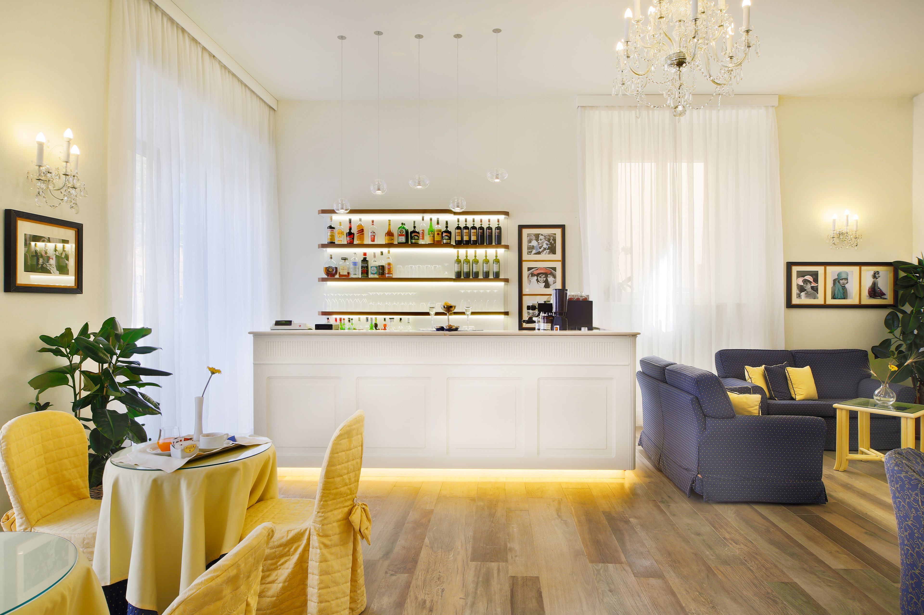 Uma área de bar com bancada branca e prateleiras cheias de garrafas, espaço de estar com sofá azul e cadeiras amarelas, e um candelabro.