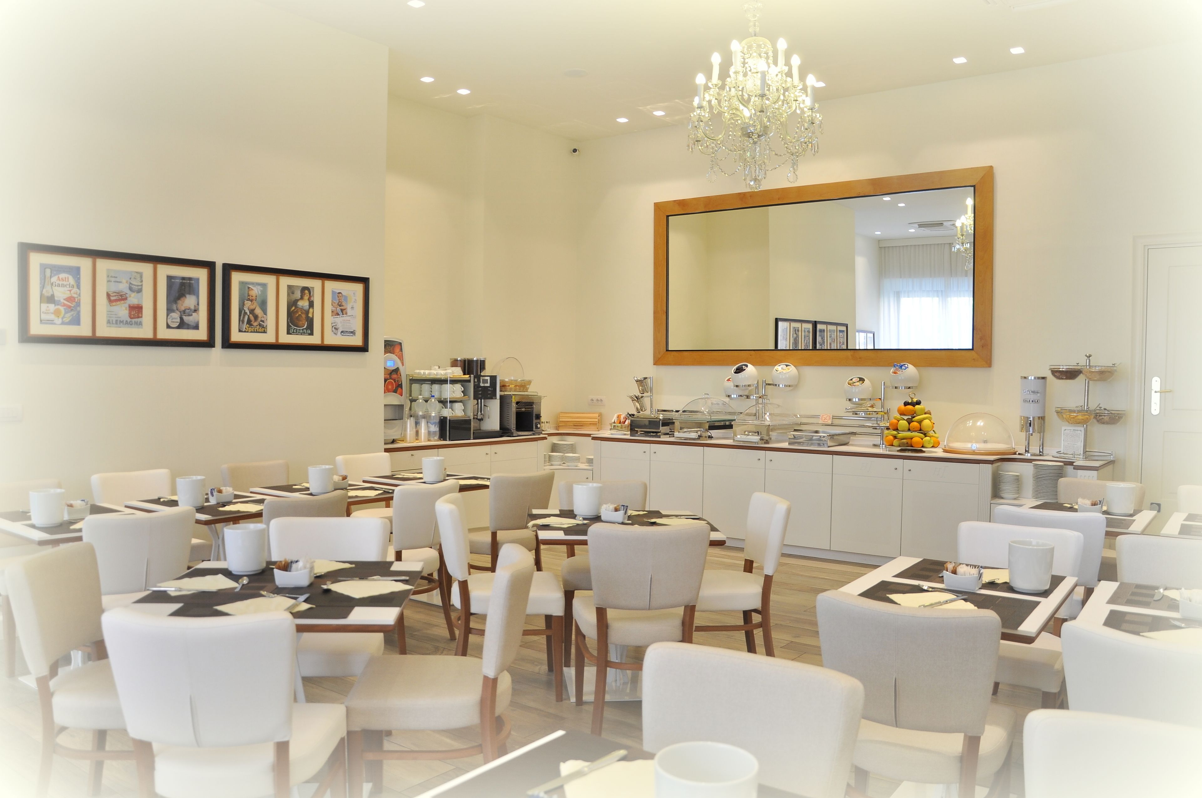 A modern hotel dining area with white chairs and tables, a central kitchen counter, and a large mirror above the counter.
