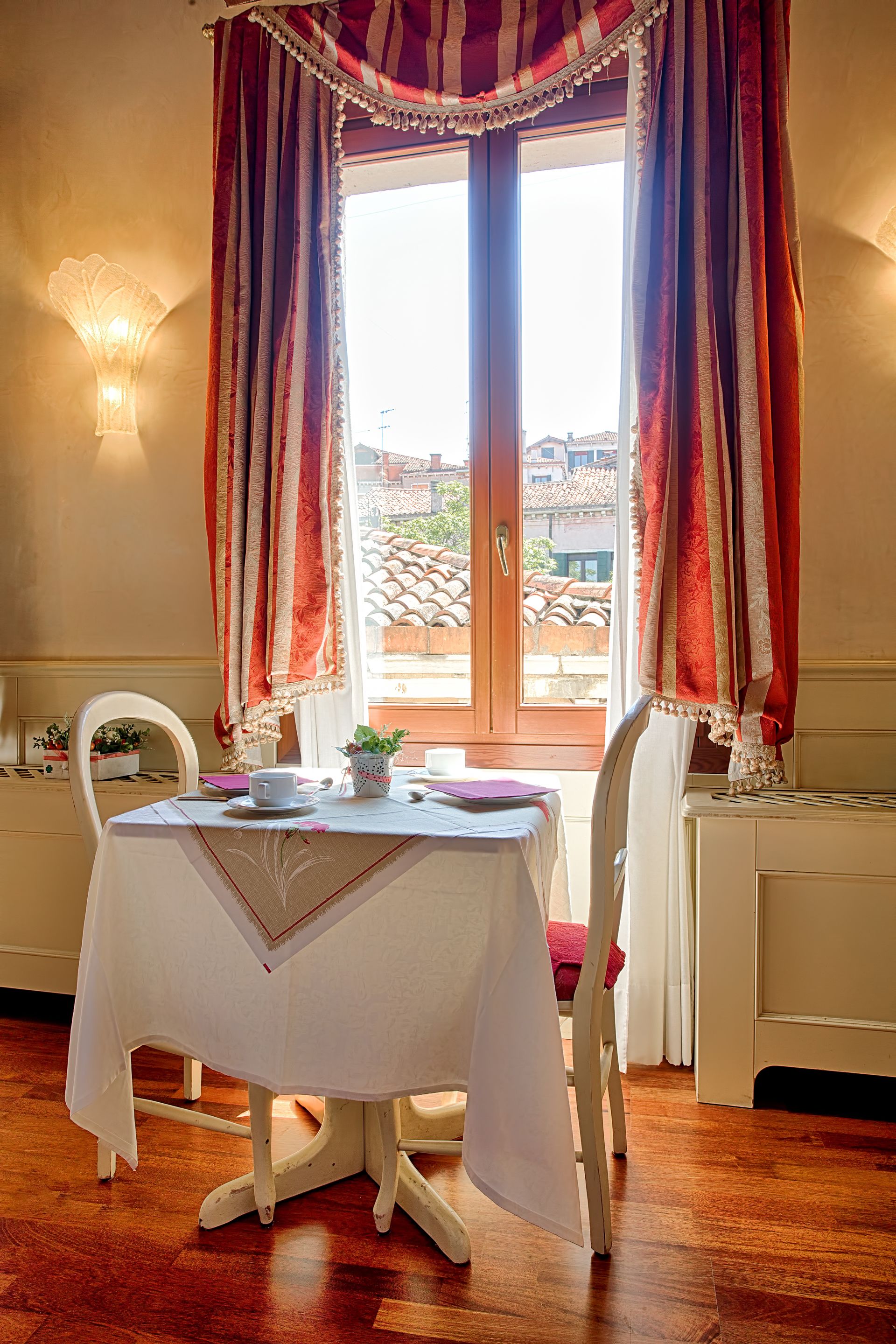 Un comedor con una mesa dispuesta para dos personas, una ventana con cortinas, una silla y una lámpara fijada a la pared.
