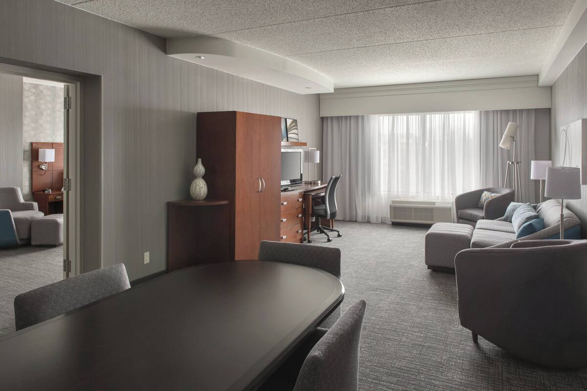 A modern hotel room with a conference table, a sofa, a desk with a computer, and a large window with curtains.