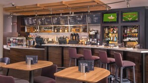 Zona de bar moderna con mesas de madera y sillas moradas, una máquina de café y un panel de menú.