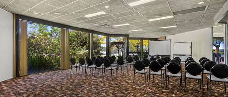 Un salón de conferencias con piso estampado, hileras de sillas, una pizarra y ventanas amplias con vista a árboles.