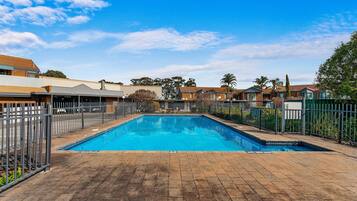 A rectangular swimming pool with a tiled deck, surrounded by a metal fence, and adjacent to residential buildings.