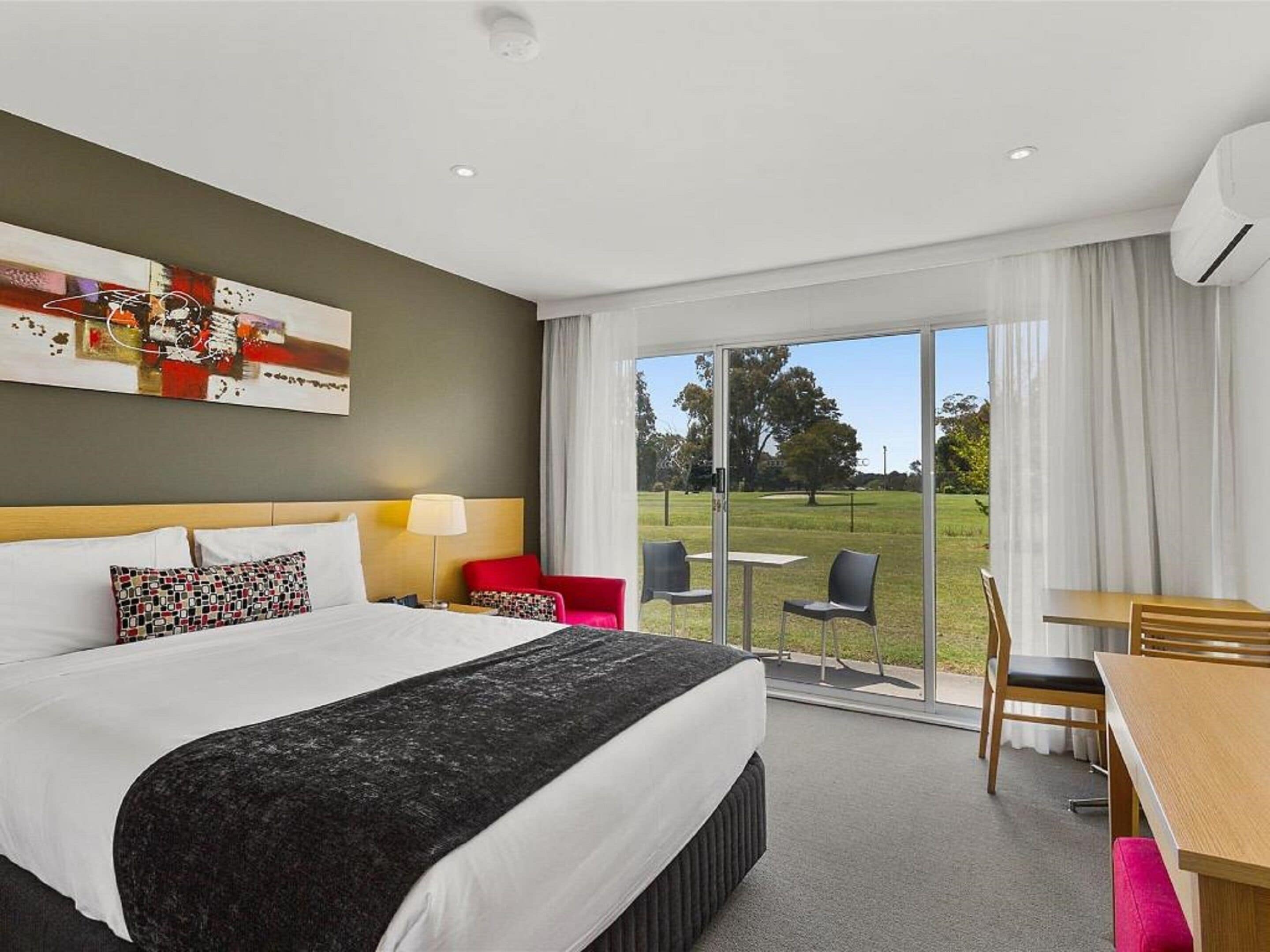 A hotel room with a bed, a desk, chairs, and a large window with a view of a grassy area.