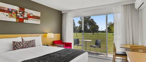 Habitación de hotel con cama, escritorio, sillas y un amplio ventanal con vistas a un área verde.