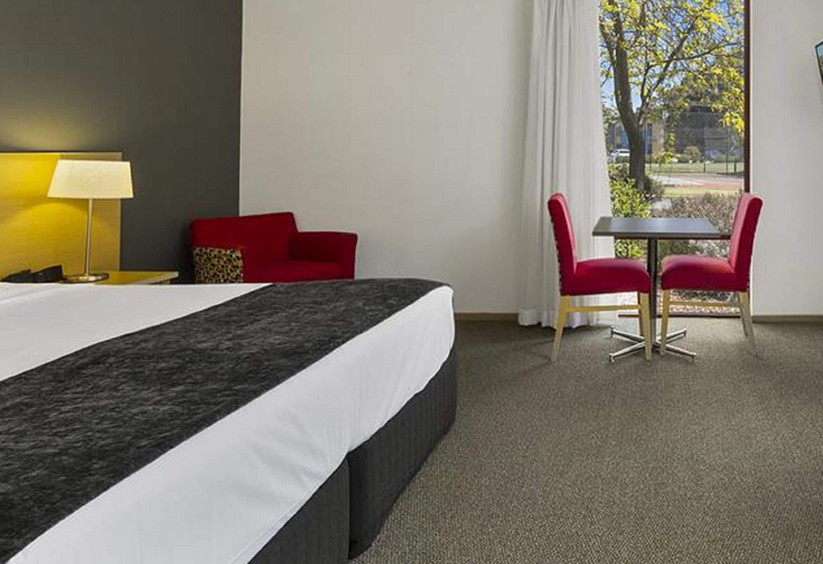 A hotel room with a large bed, a red chair, a small table, and a view of the outdoors.