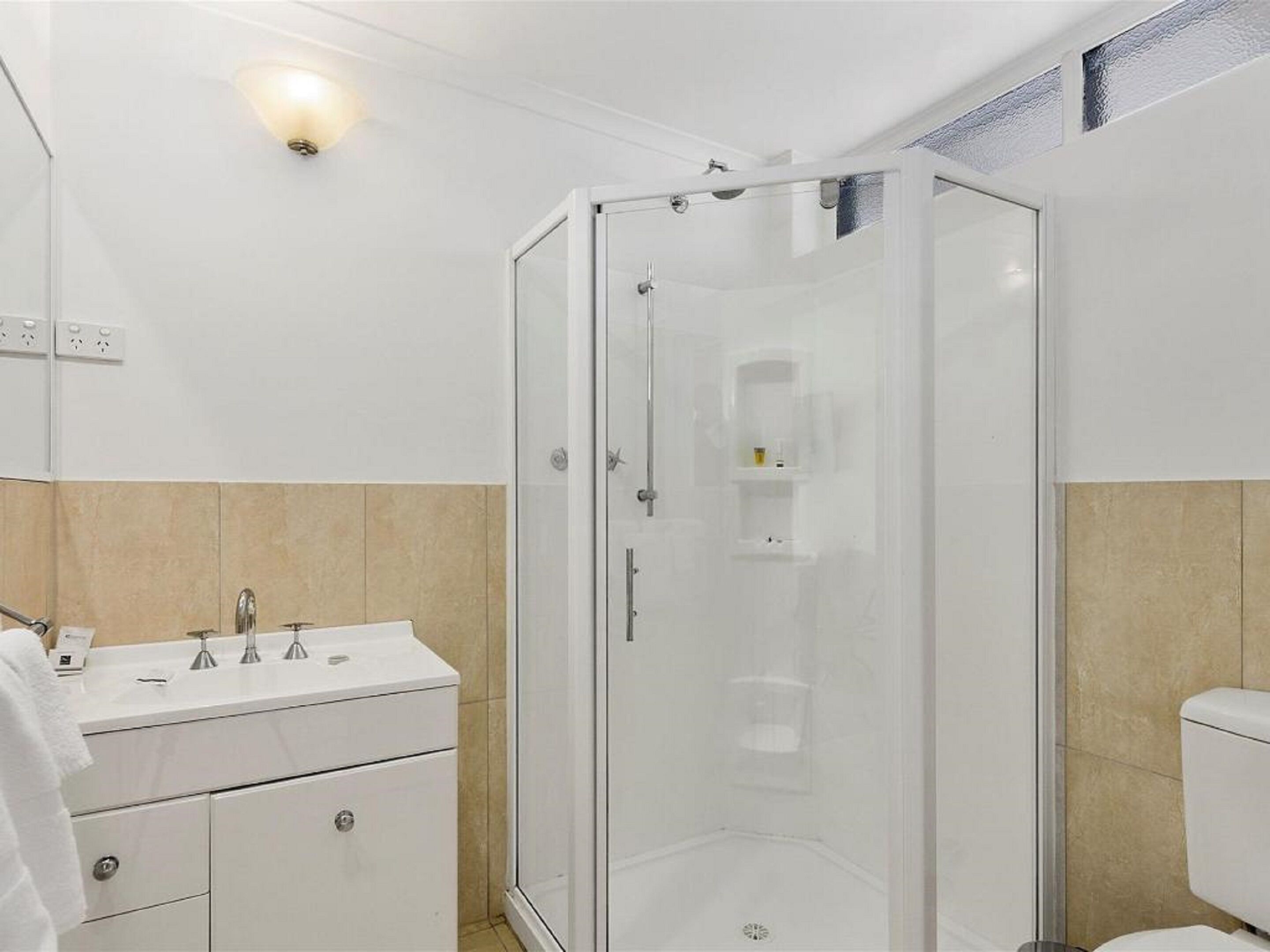 Un baño moderno con ducha de vidrio, tocador blanco con espejo y azulejos color beige.