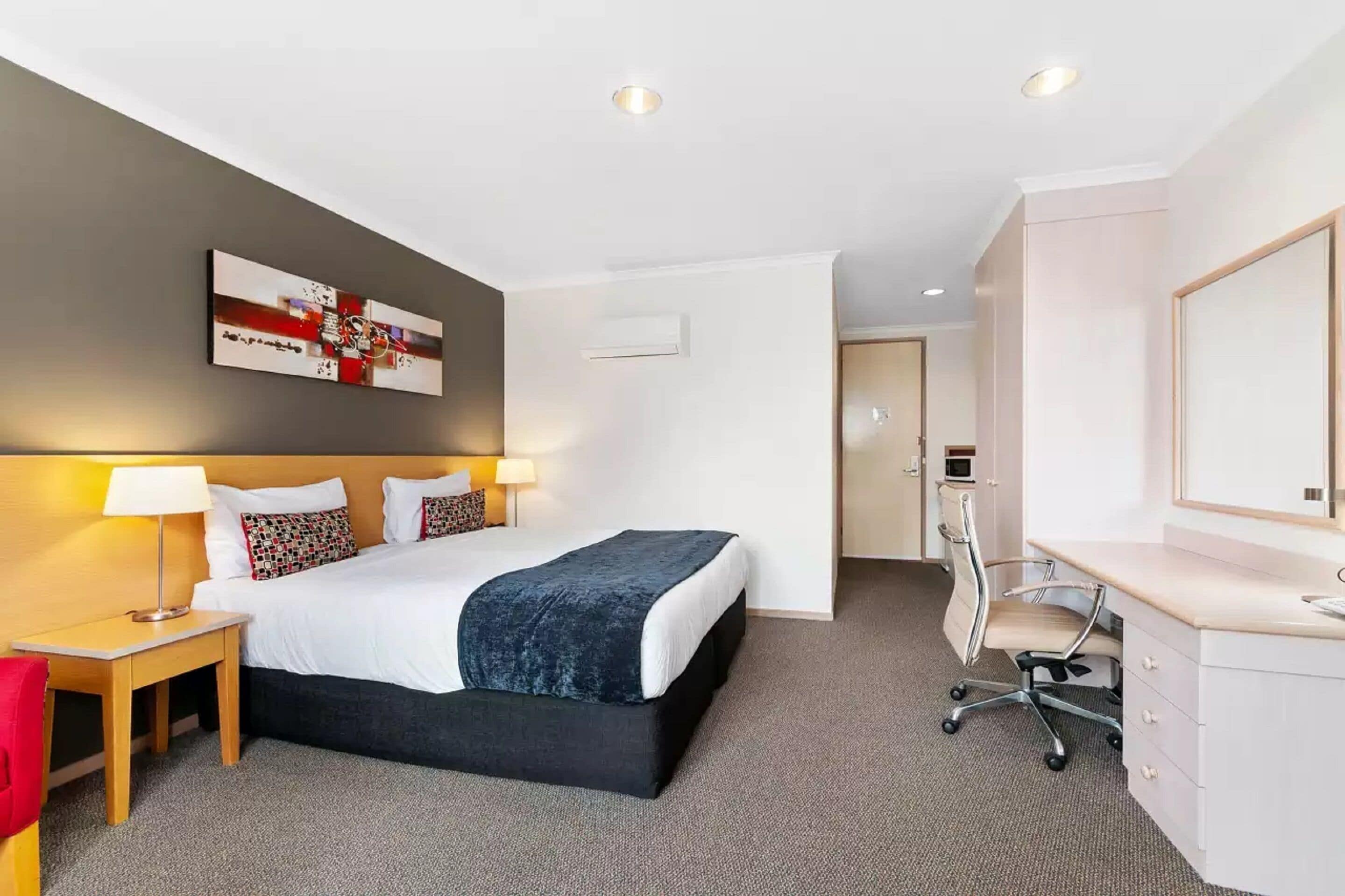 A hotel room with a bed, bedside tables, a desk, and a chair. There is a painting on the wall and an air conditioning unit on the wall.