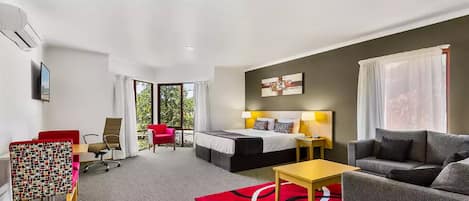 A hotel room with a bed, a sofa, a desk, and a chair. There is a window with curtains, a red rug, and an air conditioning unit on the wall.