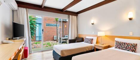 Habitación de hotel con dos camas, un escritorio y un televisor, y una puerta corrediza de cristal al exterior.