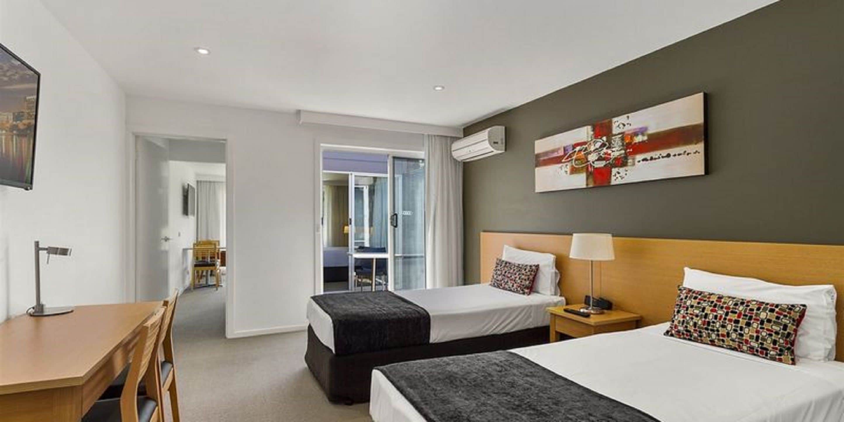 A hotel room with two beds, a desk, and a chair. There is a painting on the wall and an air conditioning unit above the bed.