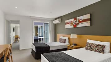 A hotel room with two beds, a desk, and a chair. There is a painting on the wall and an air conditioning unit above the bed.