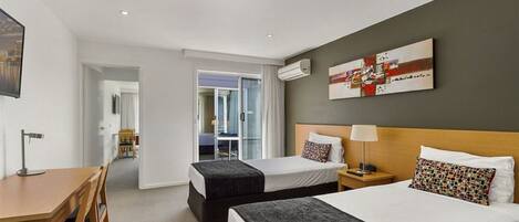 A hotel room with two beds, a desk, and a chair. There is a painting on the wall and an air conditioning unit above the bed.