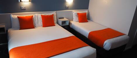Habitación de hotel con dos camas, cada una con ropa blanca y detalles naranja. Hay luces fijadas a la pared sobre las camas y una mesita de noche pequeña con una lámpara.