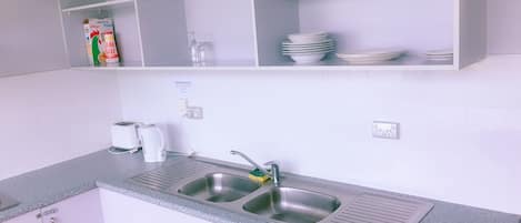 A kitchen with two stainless steel sinks, a white countertop, and a shelf above holding a box of salt, a glass, and stacked plates.