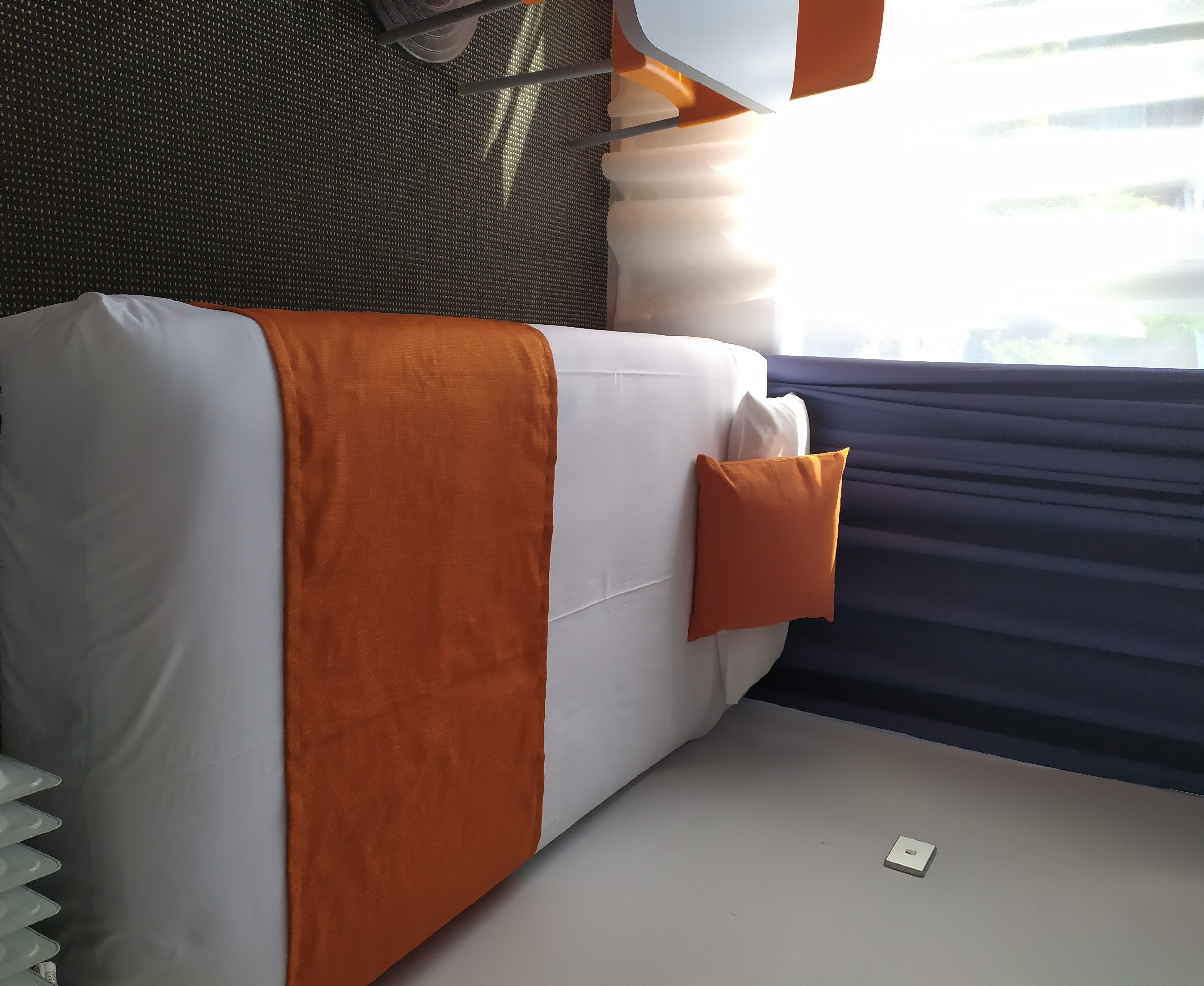 A bed with an orange blanket and pillows, a white headboard, and a window with dark curtains.