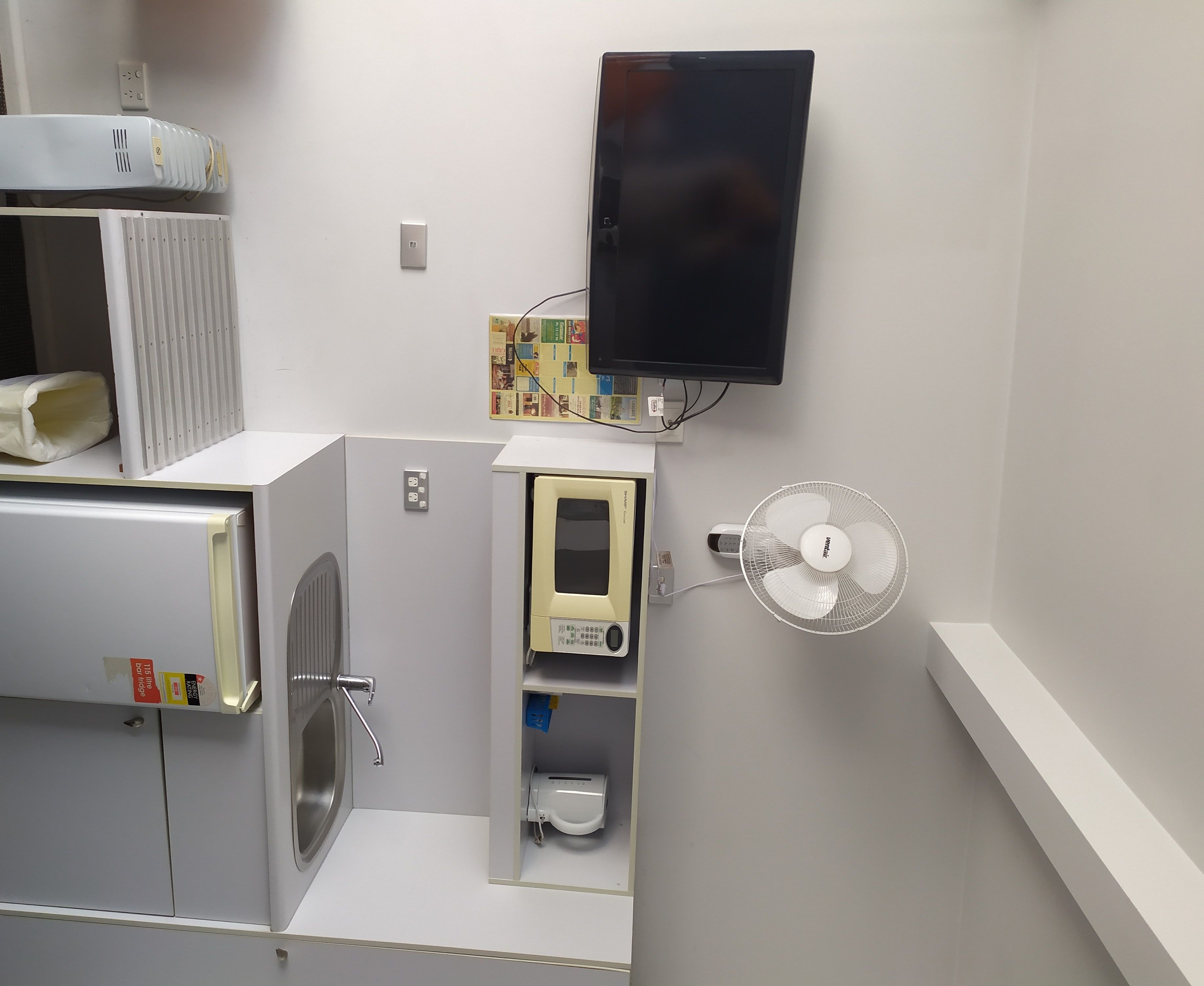 A compact kitchenette with a microwave, a small refrigerator, a built-in fan, and a wall-mounted TV.