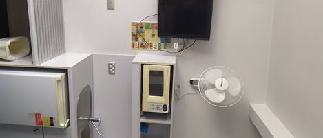 Una kitchenette compacta con microondas, un refrigerador pequeño, un ventilador integrado y un televisor montado en la pared.