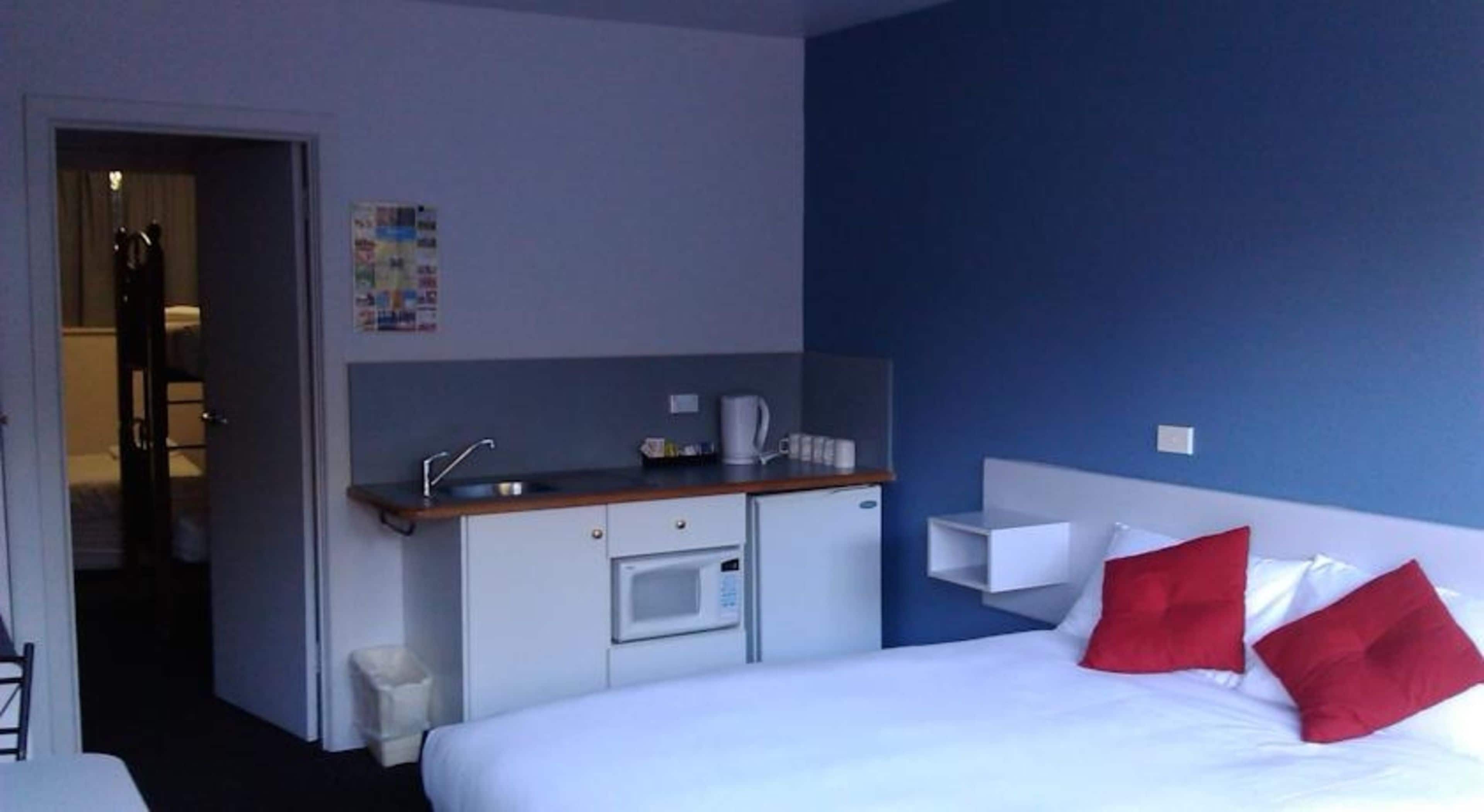 A hotel room with a bed, a small kitchenette, a white trash bin, and a door leading to another room.