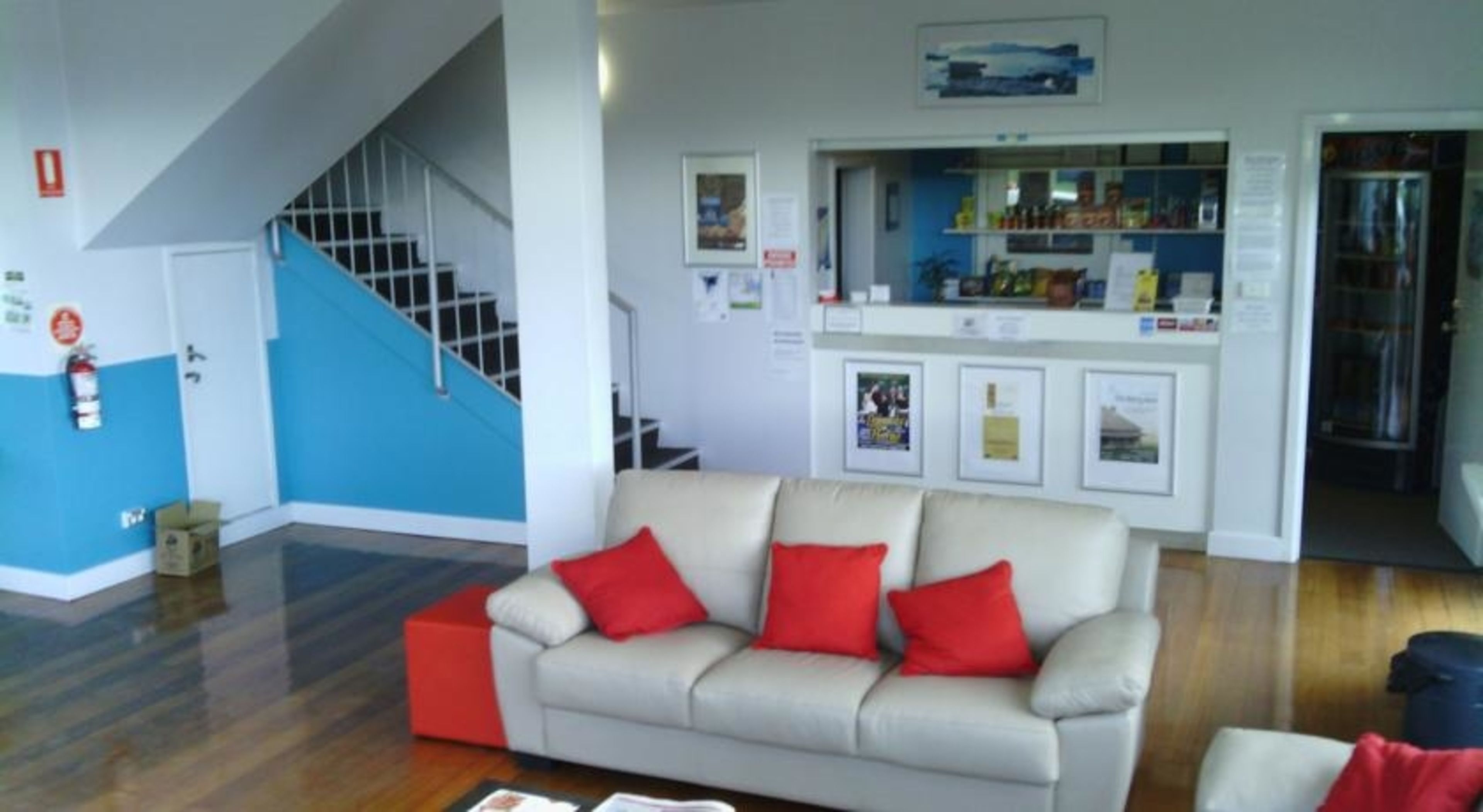 A living room with a beige sofa, red cushions, a staircase, and a kitchen area with a counter and various items.