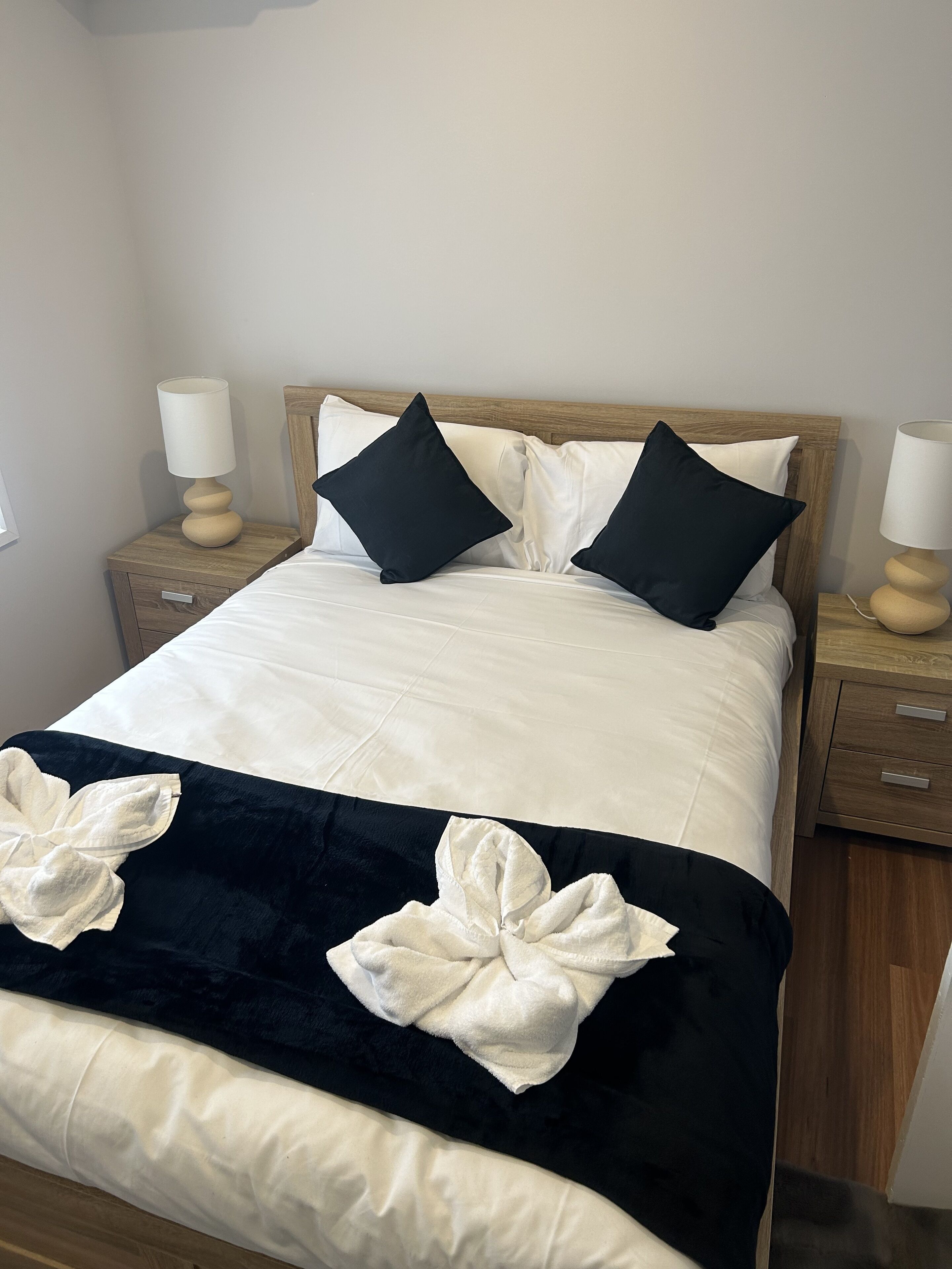 A neatly made bed with a white and black bedspread, two black pillows, and a wooden headboard. There are two bedside tables with lamps and a small drawer.