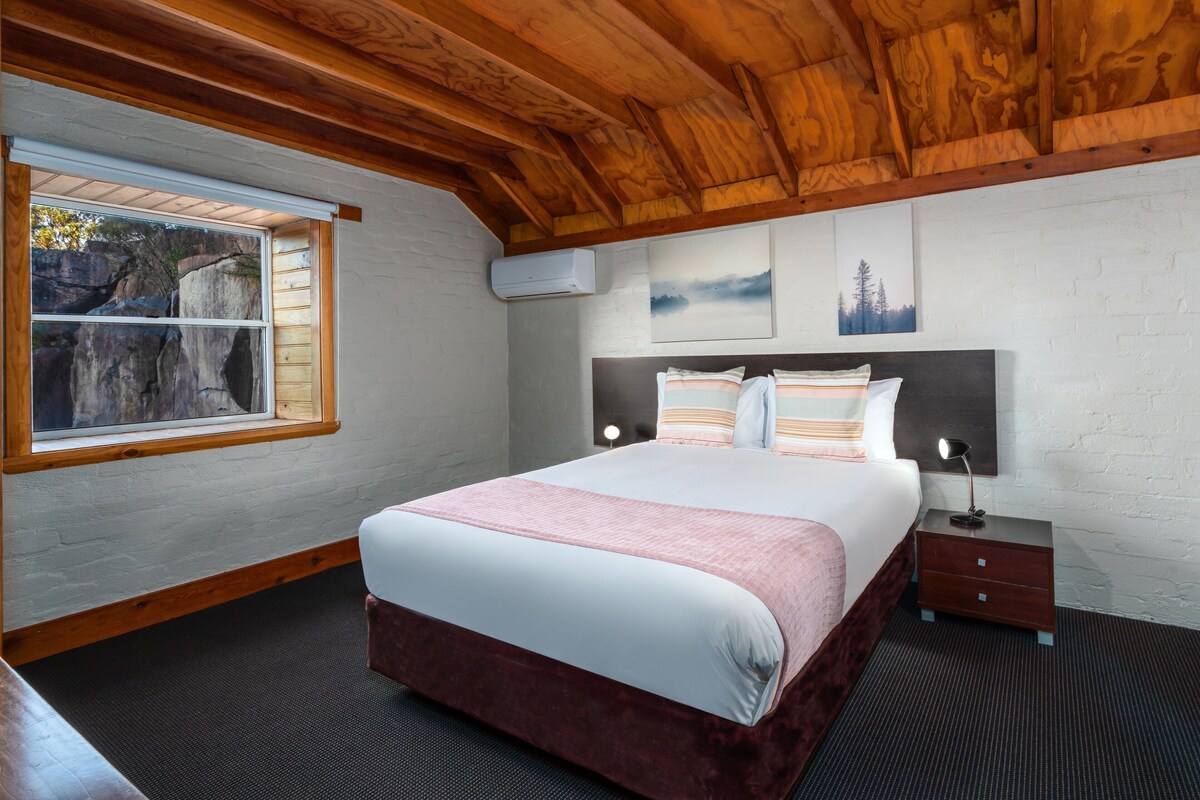 A bedroom with a large bed, a nightstand, a window with a view of rocks, and wooden ceiling beams.