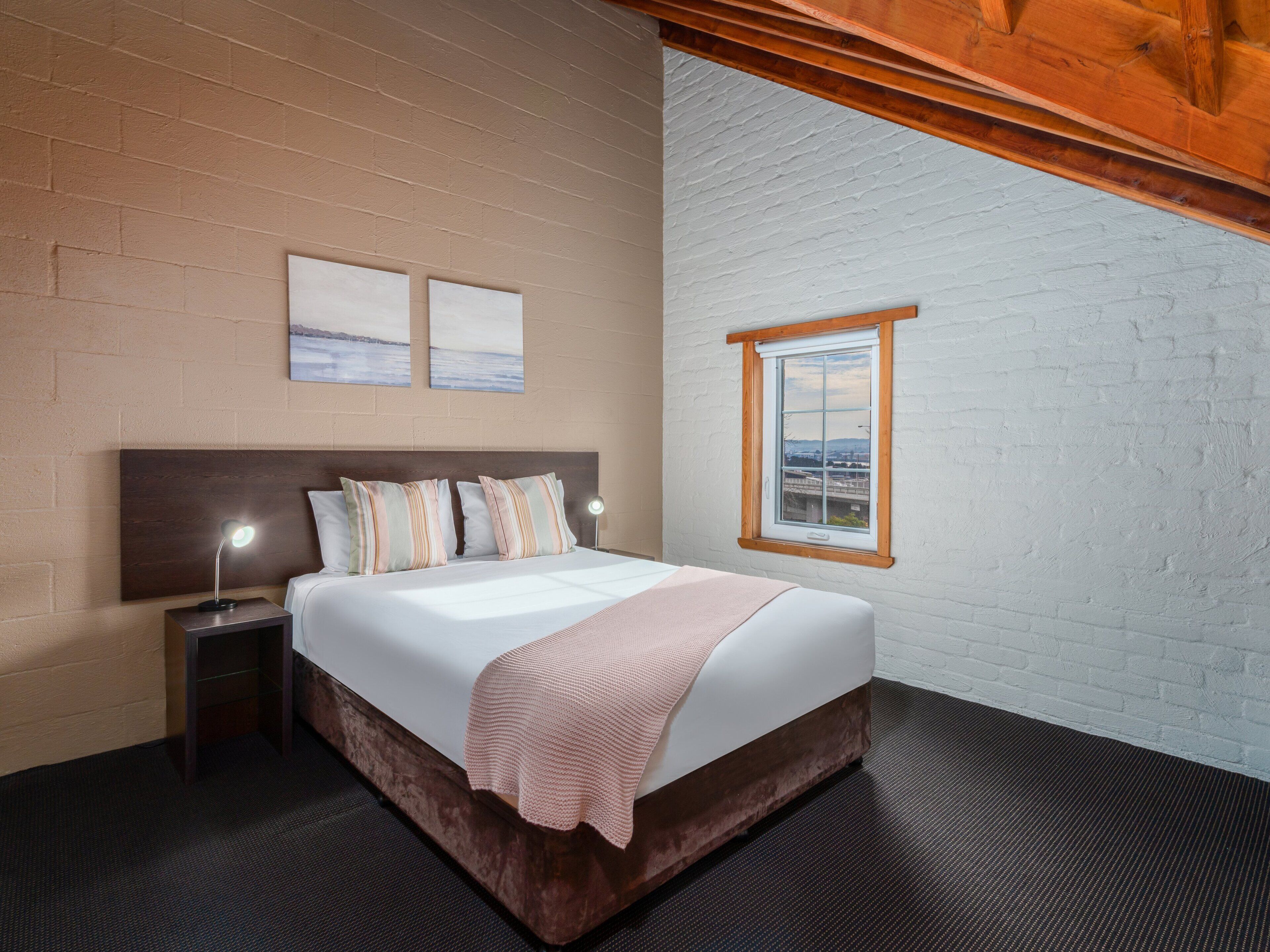 A bedroom with a large bed, a nightstand, a lamp, two wall art pieces, and a window with a view.