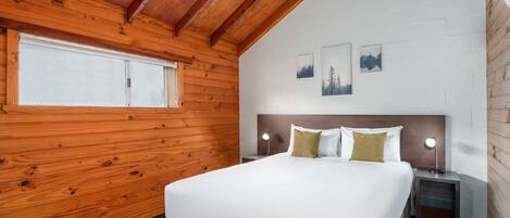 Un dormitorio con techo de madera, una cama con ropa de cama blanca, dos mesitas de noche con lámparas y tres cuadros enmarcados en la pared.