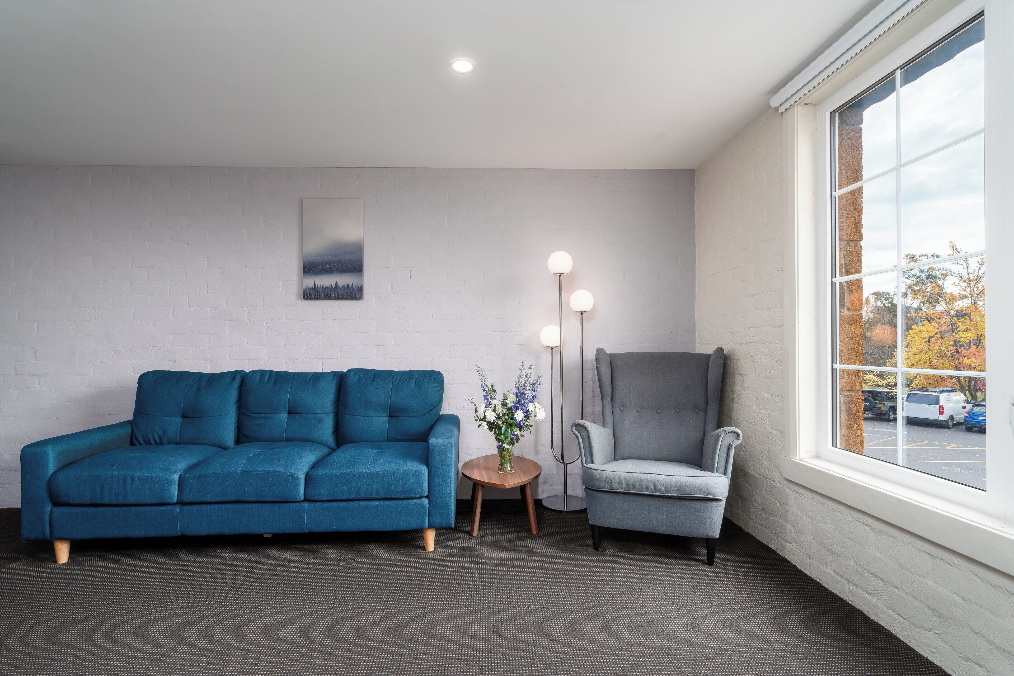 A modern room with a blue sofa, a grey armchair, a small round table with a vase, and a large window with a view of the outside.