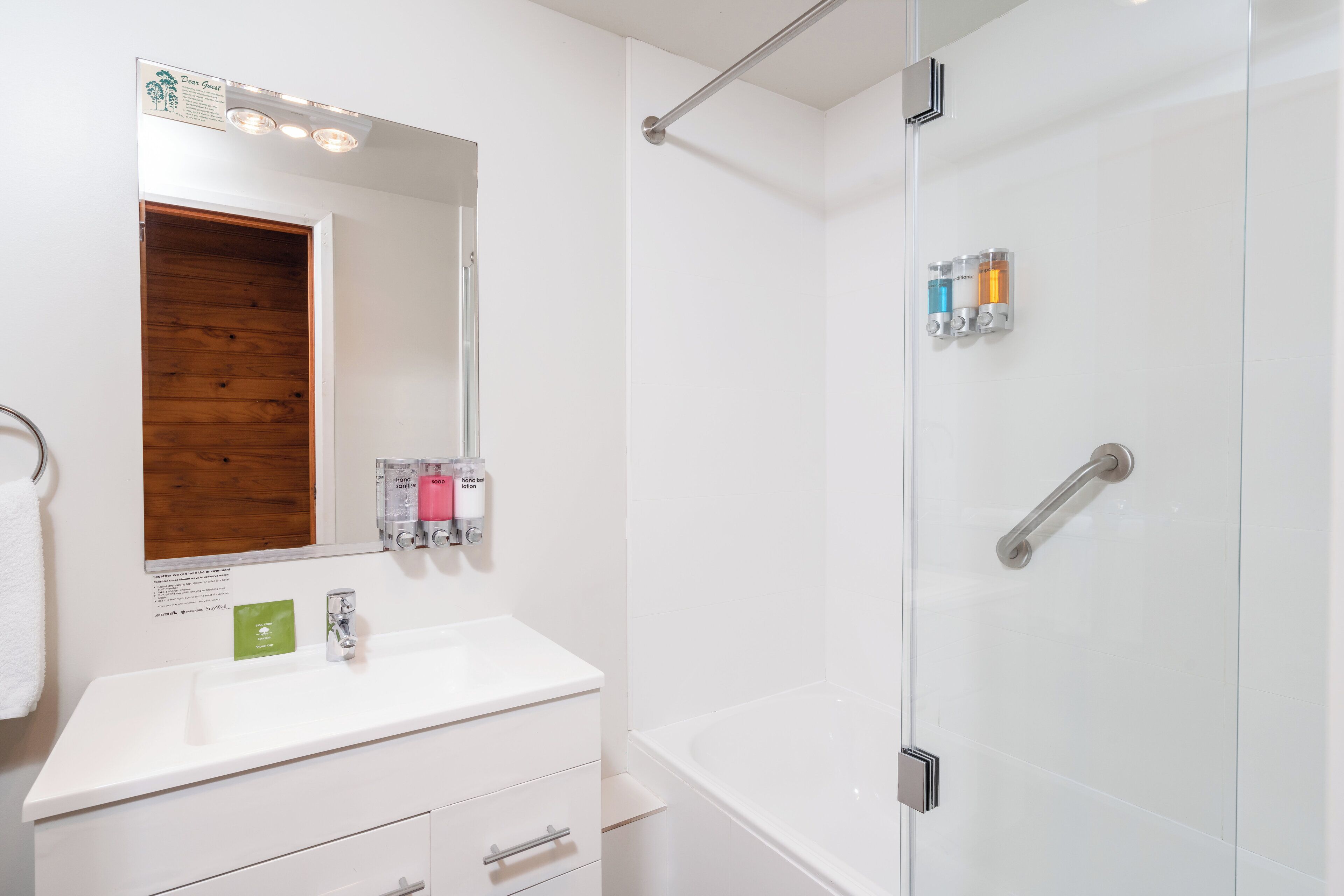 Un baño moderno con un tocador blanco, un espejo grande, un box de ducha de vidrio y un panel de madera sobre el espejo.