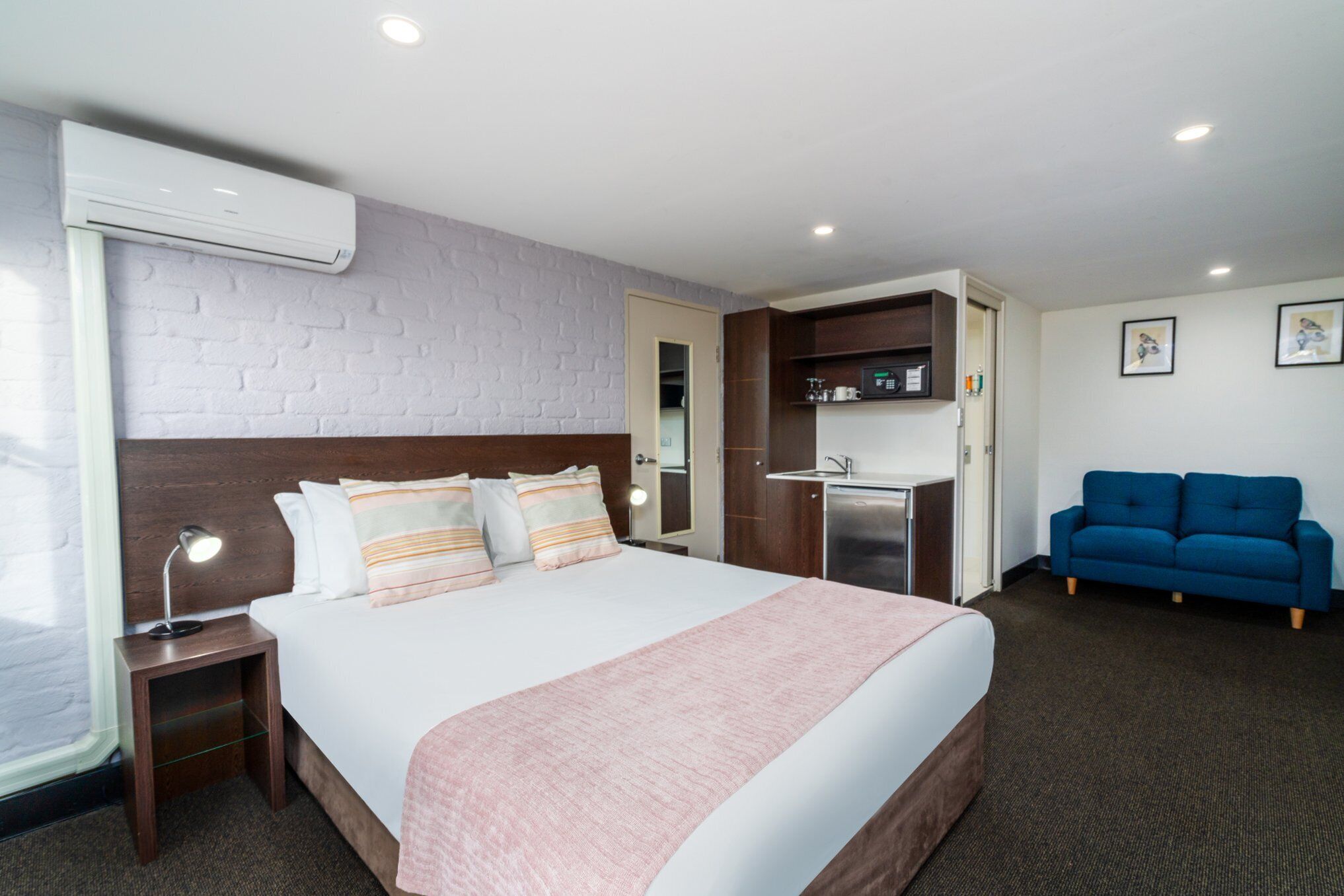 A hotel room with a bed, bedside table, lamp, air conditioning unit, kitchenette, and a blue sofa.