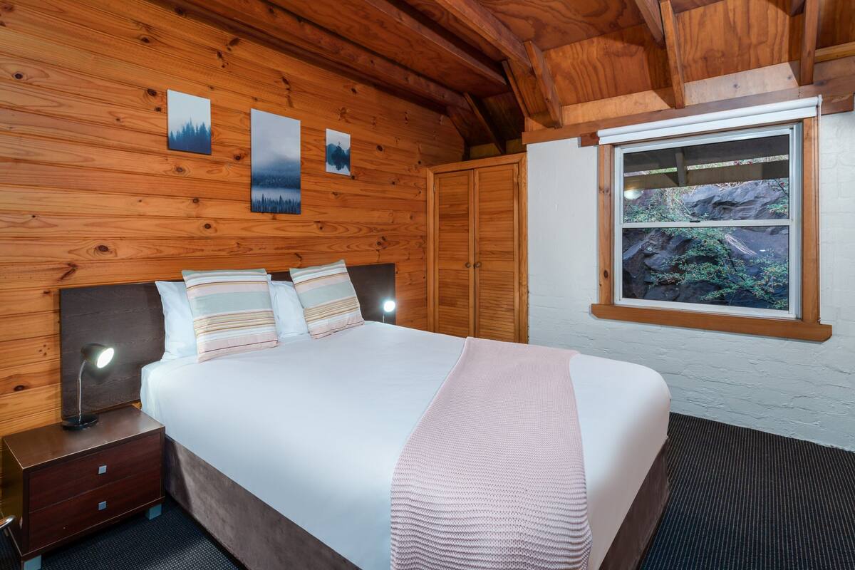 A wooden-paneled bedroom with a bed, nightstands, and a window with a view of rocks and greenery.