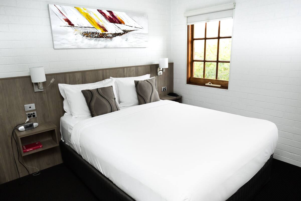 A neatly made bed in a hotel room with a white headboard, a bedside table with a lamp, and a window with a view of greenery.