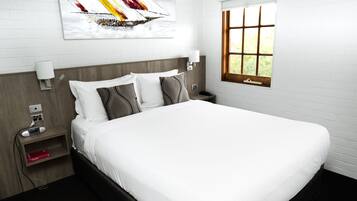 A neatly made bed in a hotel room with a white headboard, a bedside table with a lamp, and a window with a view of greenery.