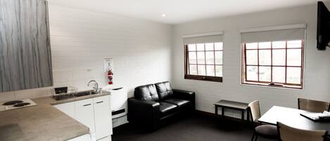 A modern kitchen with white cabinets, a black leather sofa, a dining table with chairs, and large windows with blinds.