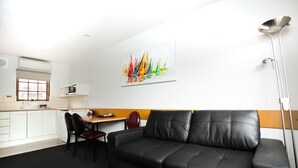 A modern hotel room with a black leather sofa, a dining table with chairs, a kitchen area, and a colorful abstract painting on the wall. - Bay Hotel Apartments (Sandy Bay)