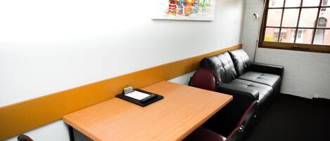 A small, private room with a wooden table, a black leather sofa, and a single red chair. A colorful abstract painting is on the wall, and a window with blinds is visible.