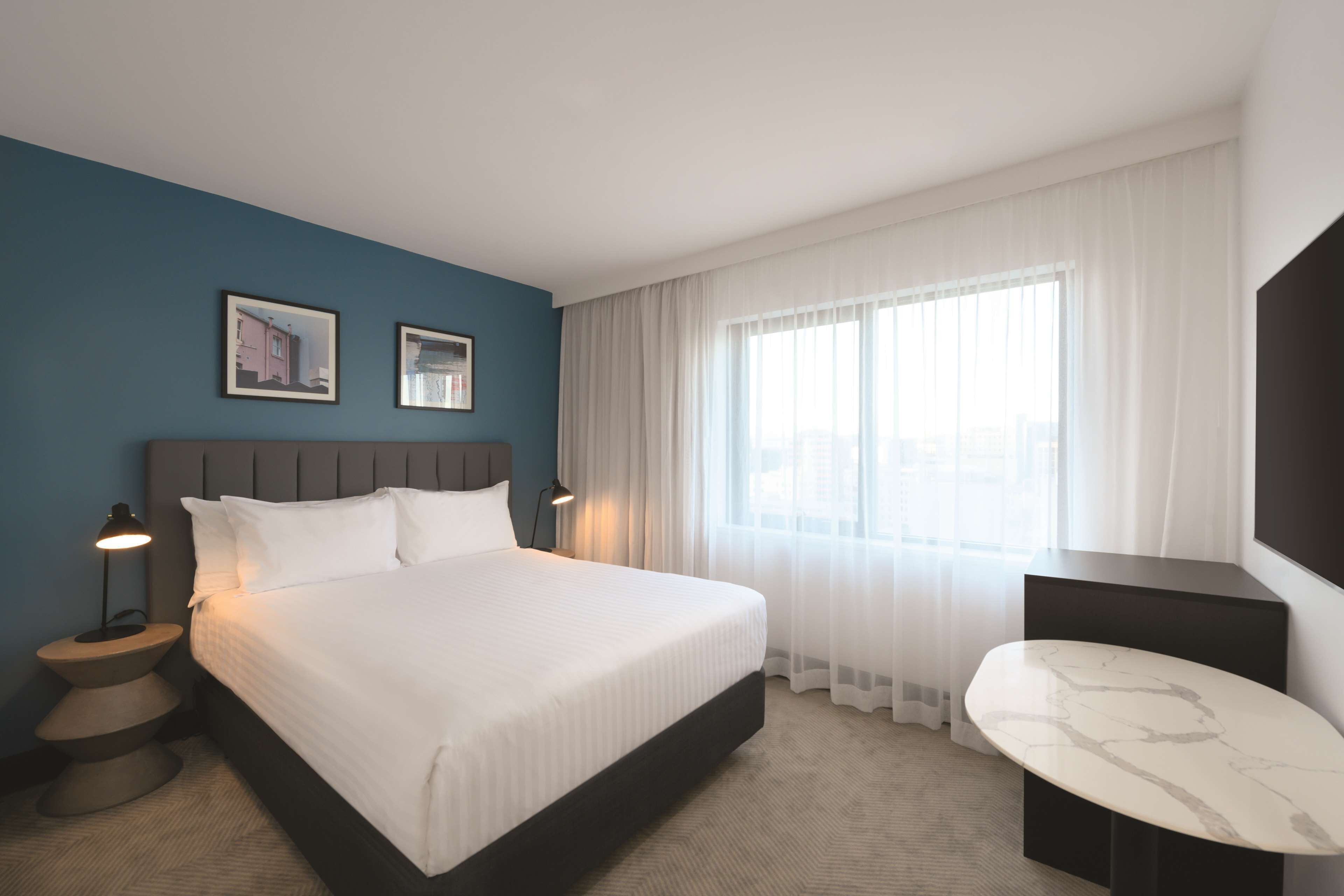 A hotel room with a large bed, a bedside table with a lamp, a small table with a marble top, and a large window with sheer curtains.