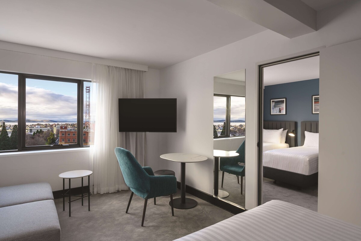 A hotel room with a large window, a flat-screen TV, a small round table, a teal chair, a bed, and a mirror reflecting the room.
