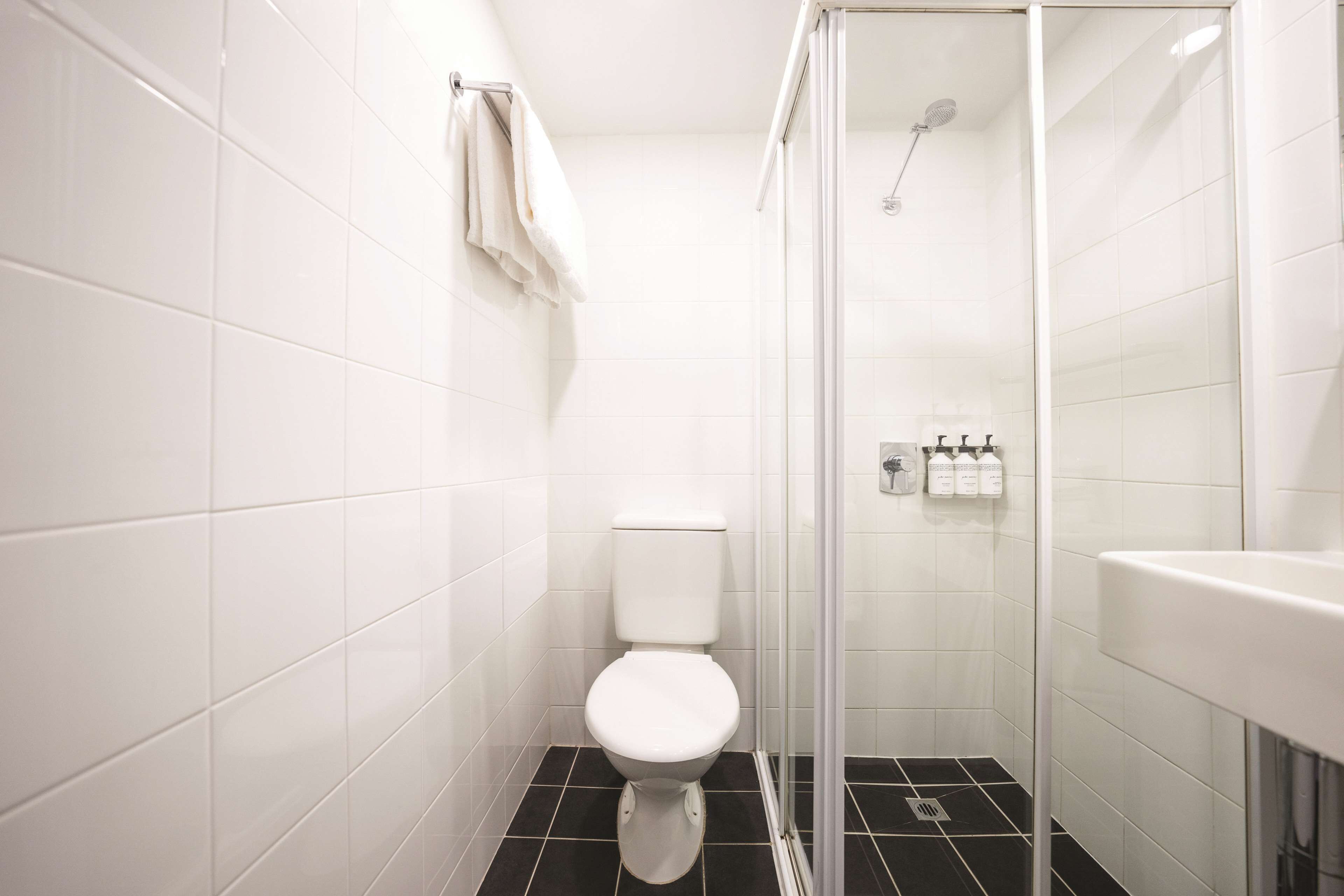 A small, modern bathroom with white tiles, a white toilet, a glass shower enclosure, and a white sink.