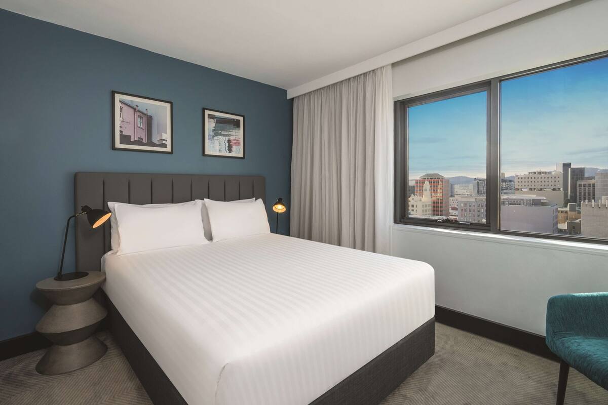 Ein Hotelzimmer mit einem großen Bett, einem Nachttisch mit Lampe, einem Sessel und Blick auf die Stadt durch ein großes Fenster.