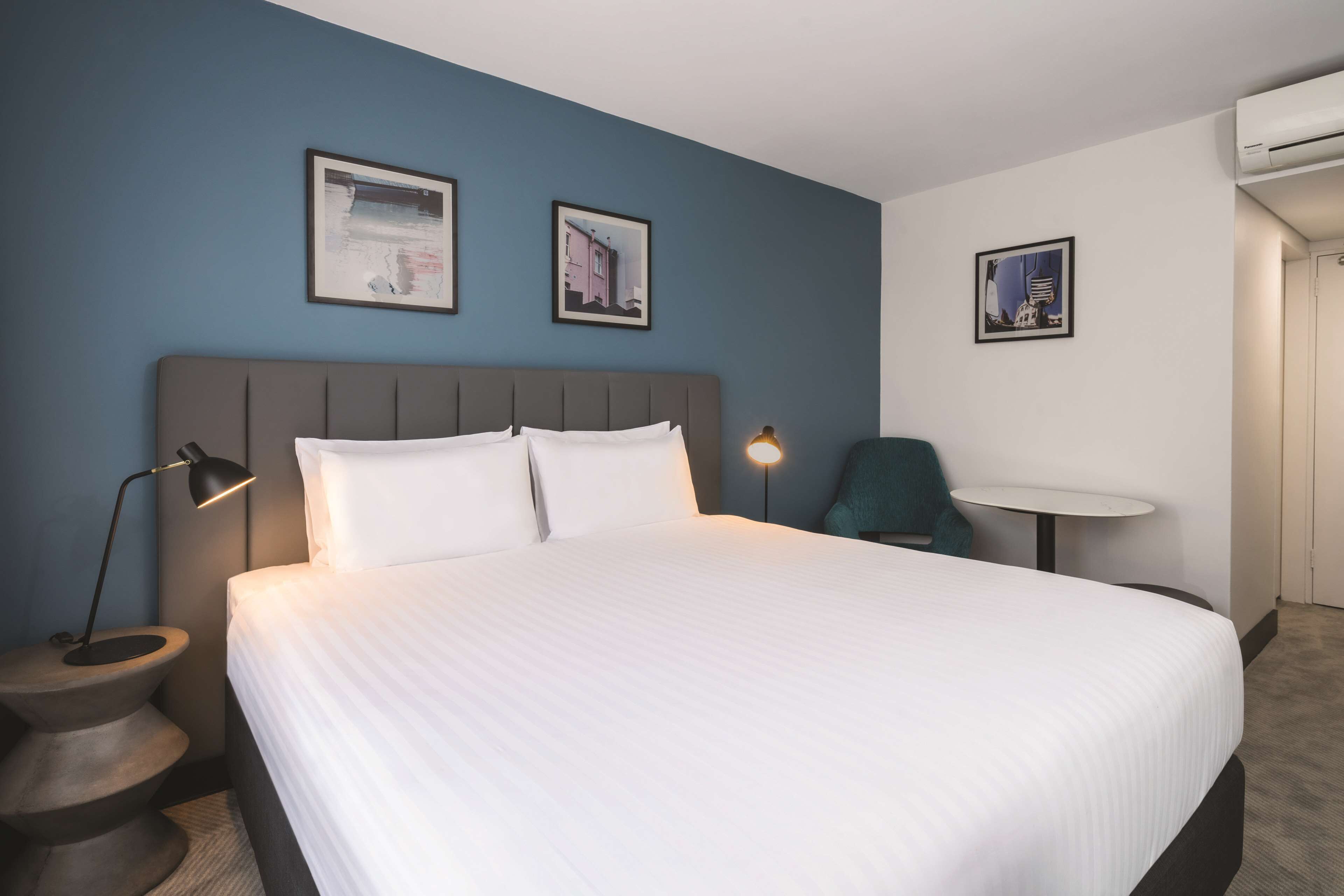 Ein Hotelzimmer mit einem großen Bett, einer Nachttischlampe, einem kleinen runden Tisch und zwei gerahmten Bildern an der Wand.