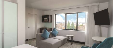 Habitación de hotel moderna con una cama, un sofá con dos almohadas, una mesita redonda pequeña, un televisor montado en la pared y una ventana con vistas a la ciudad.