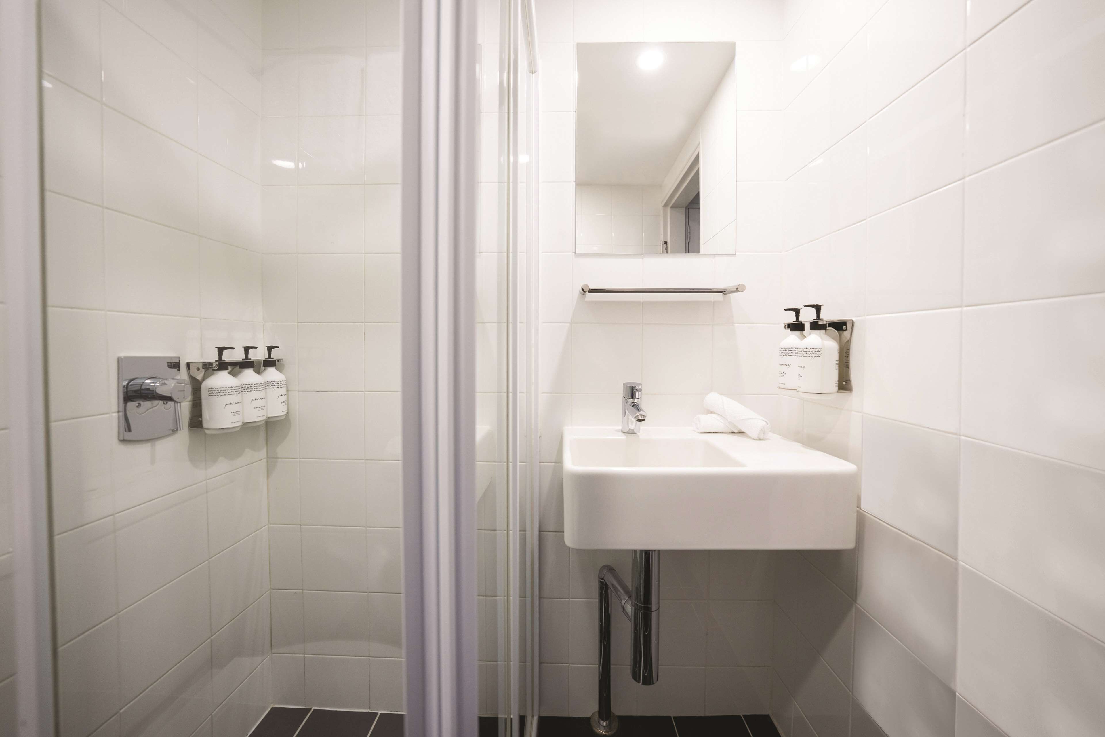 Un baño moderno con azulejos blancos, cabina de ducha con mampara de vidrio, lavamanos con espejo y artículos de aseo en la pared.